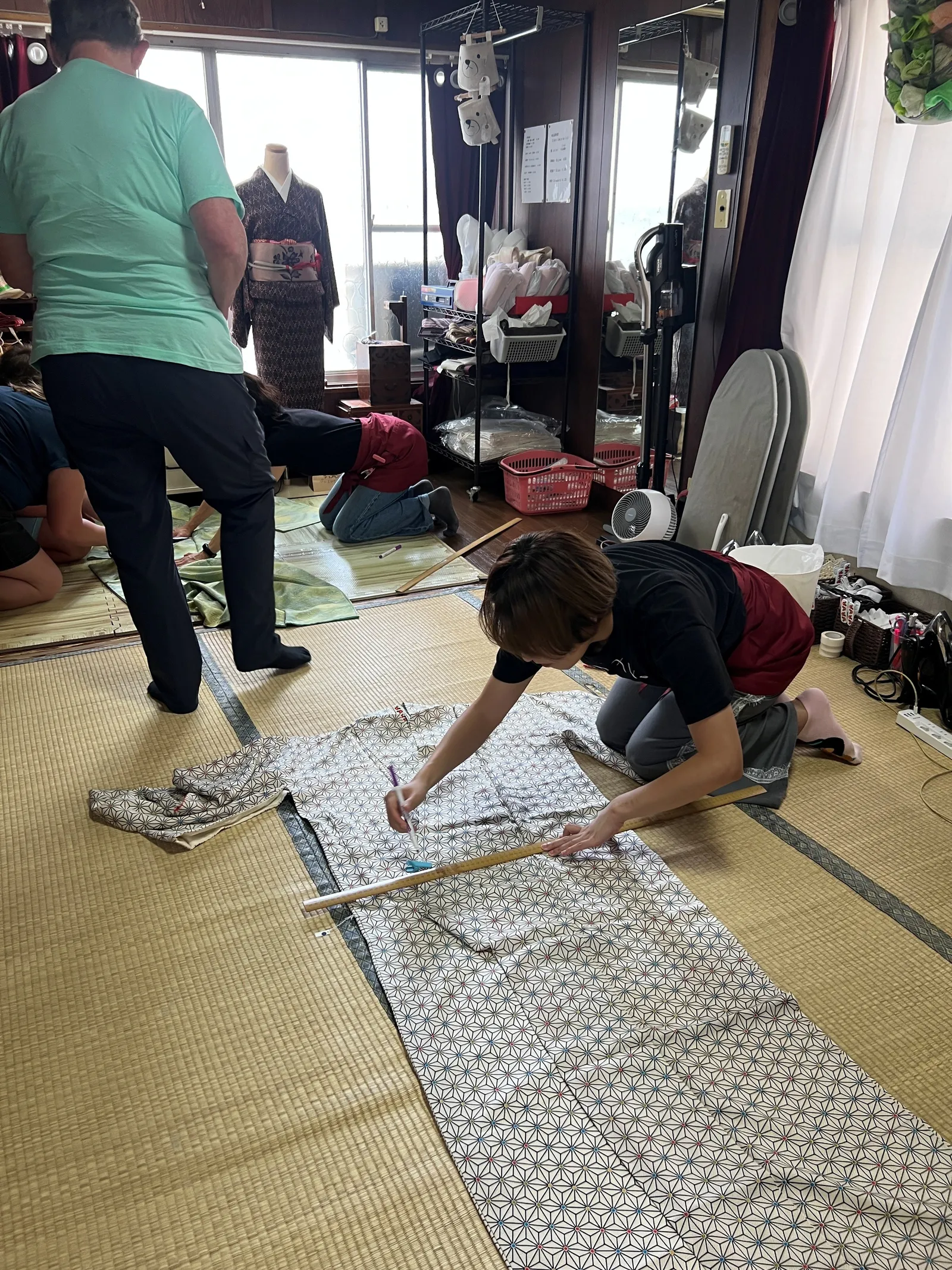 畳の上で着物の生地に定規を当てて印を付けるスタッフと、奥で作業する参加者