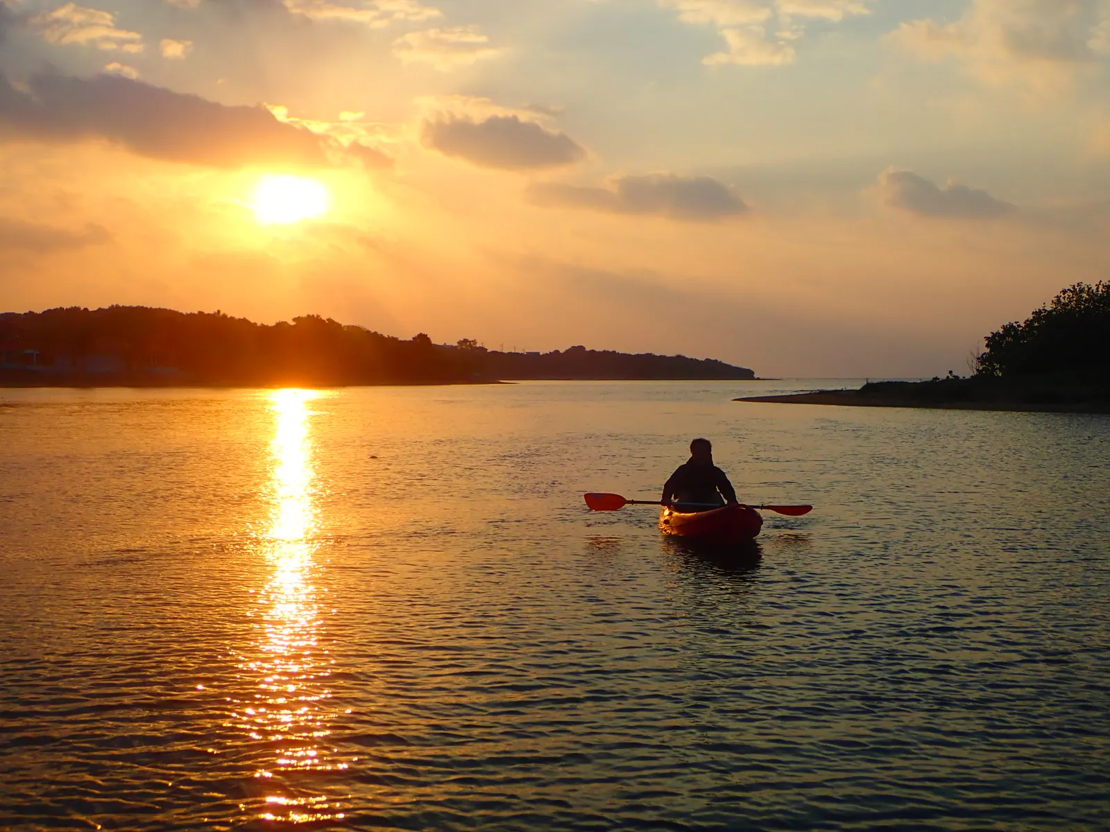 The back of a person paddling a red canoe toward a sunrise that looks like a sunset