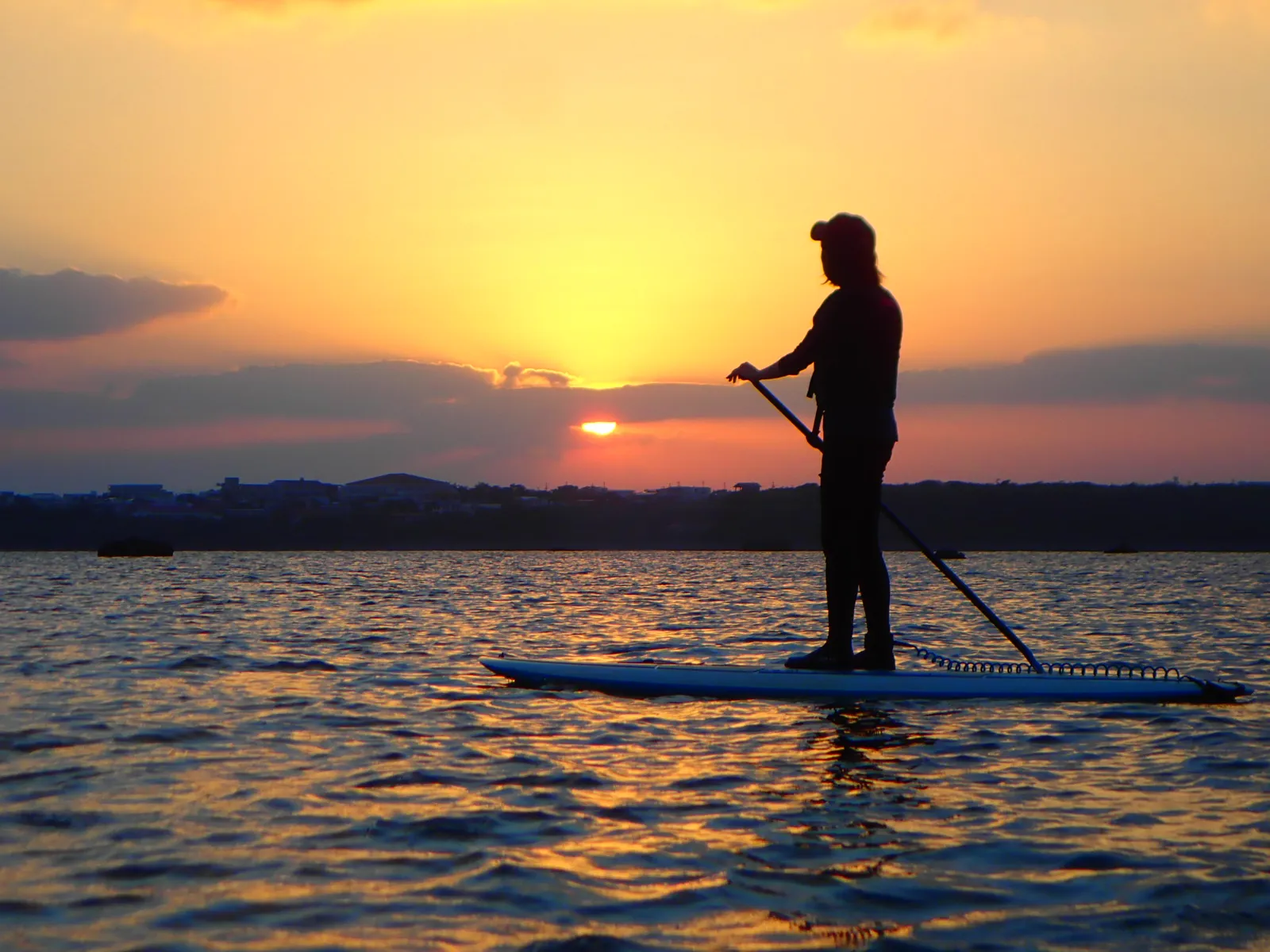 街並みの向こうに沈む夕日とSUPに立つ人のシルエット