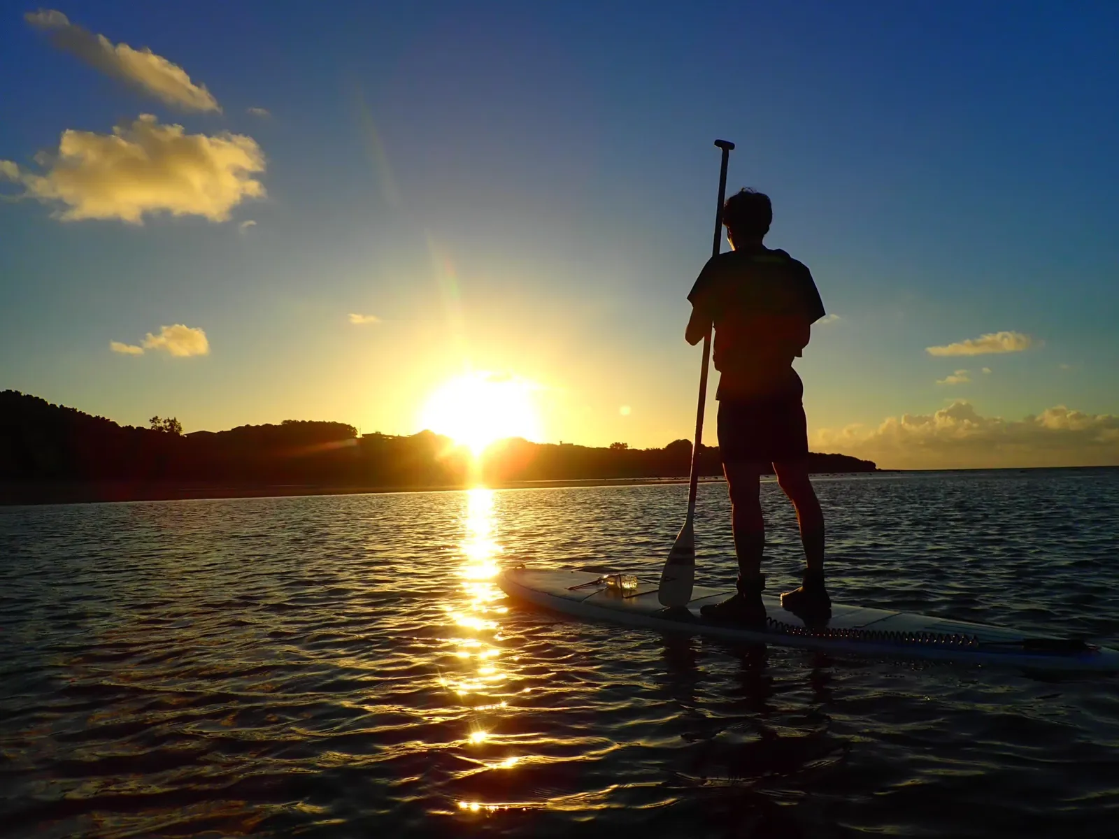 The back of a person standing on a SUP beside the rising sun