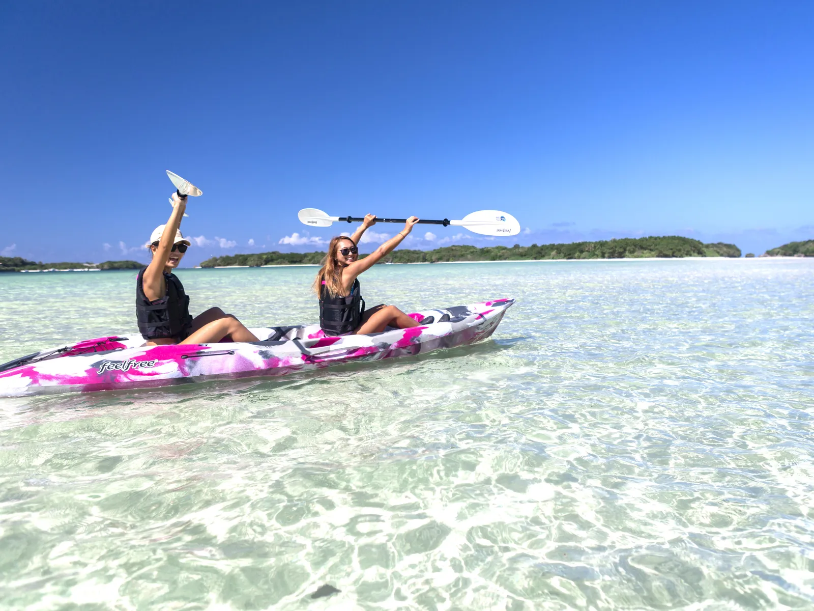 Women in a two-person canoe raising their paddles in clear shallow water