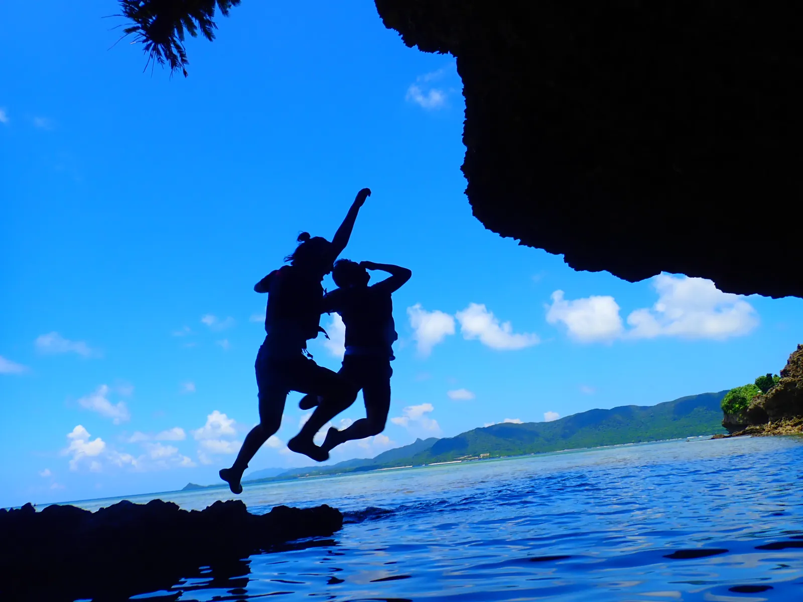 Silhouettes of two people jumping, shot from the shade of the cave rocks against the sea and blue sky