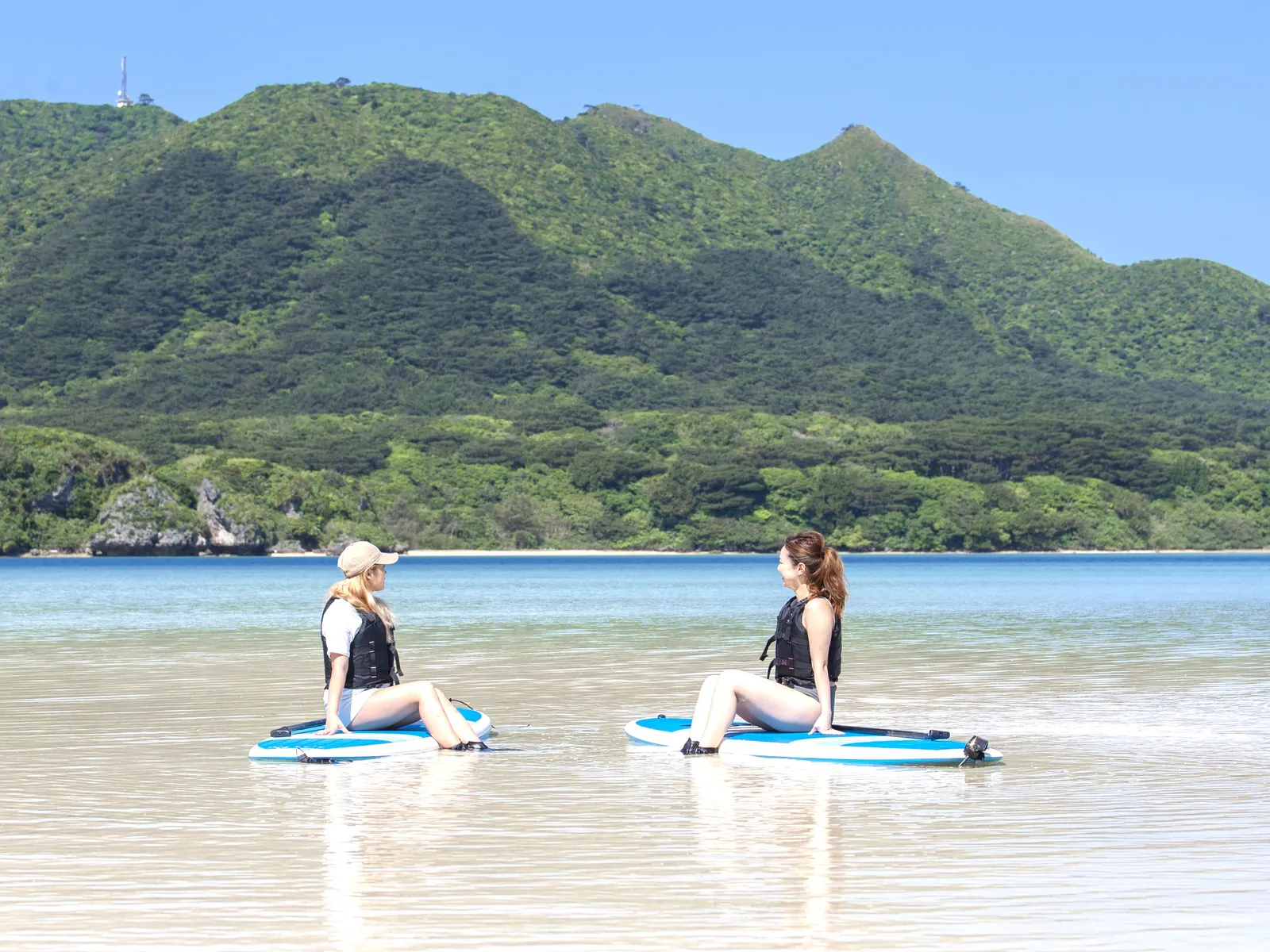 Two women sitting facing each other on SUP boards with green mountains behind them
