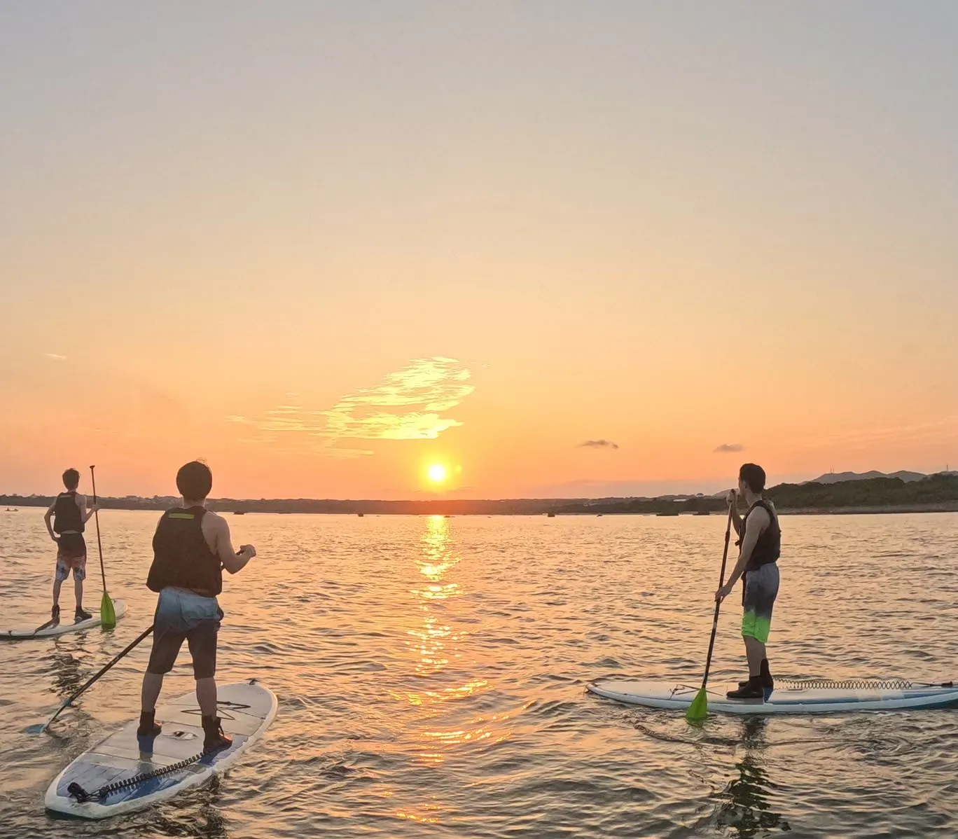 ライフジャケットを着てSUPボードに立ち、夕日の方向を見る3人