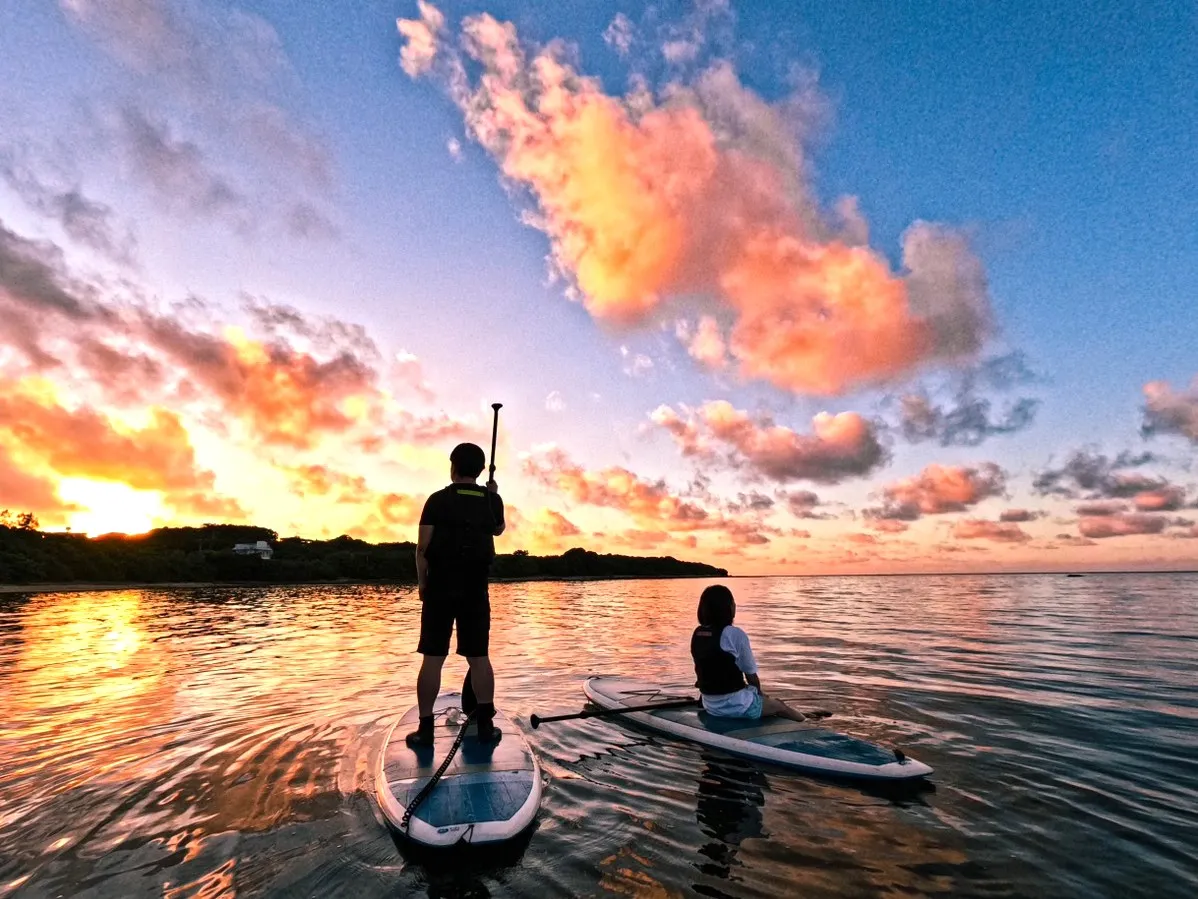 One person standing and another sitting on SUPs under pink clouds