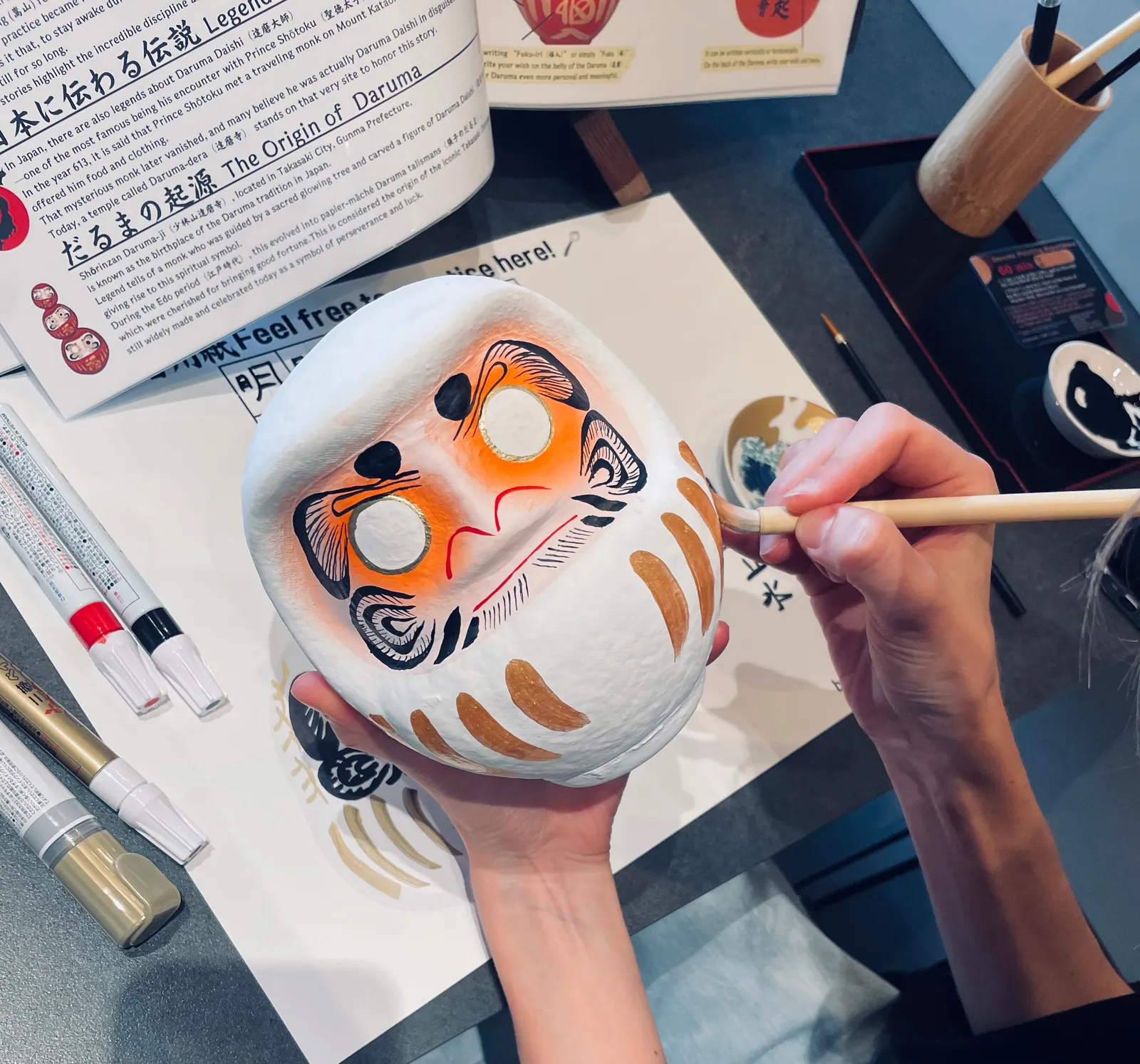 白いだるまを手に持ち、筆で金色の模様を描き入れる手元