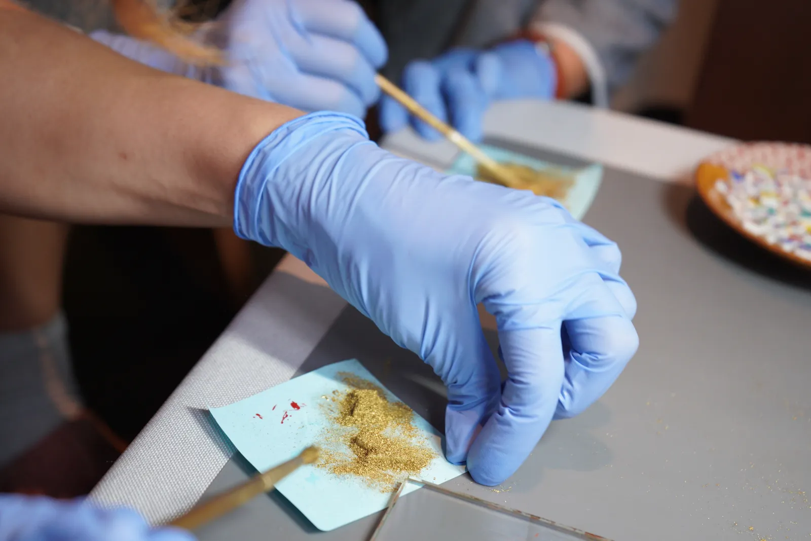 A hand in a blue glove handling gold powder with a brush