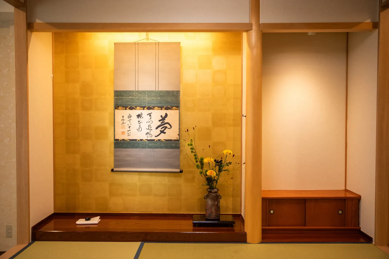 金色の壁の床の間に掛け軸と黄色い花の生け花
