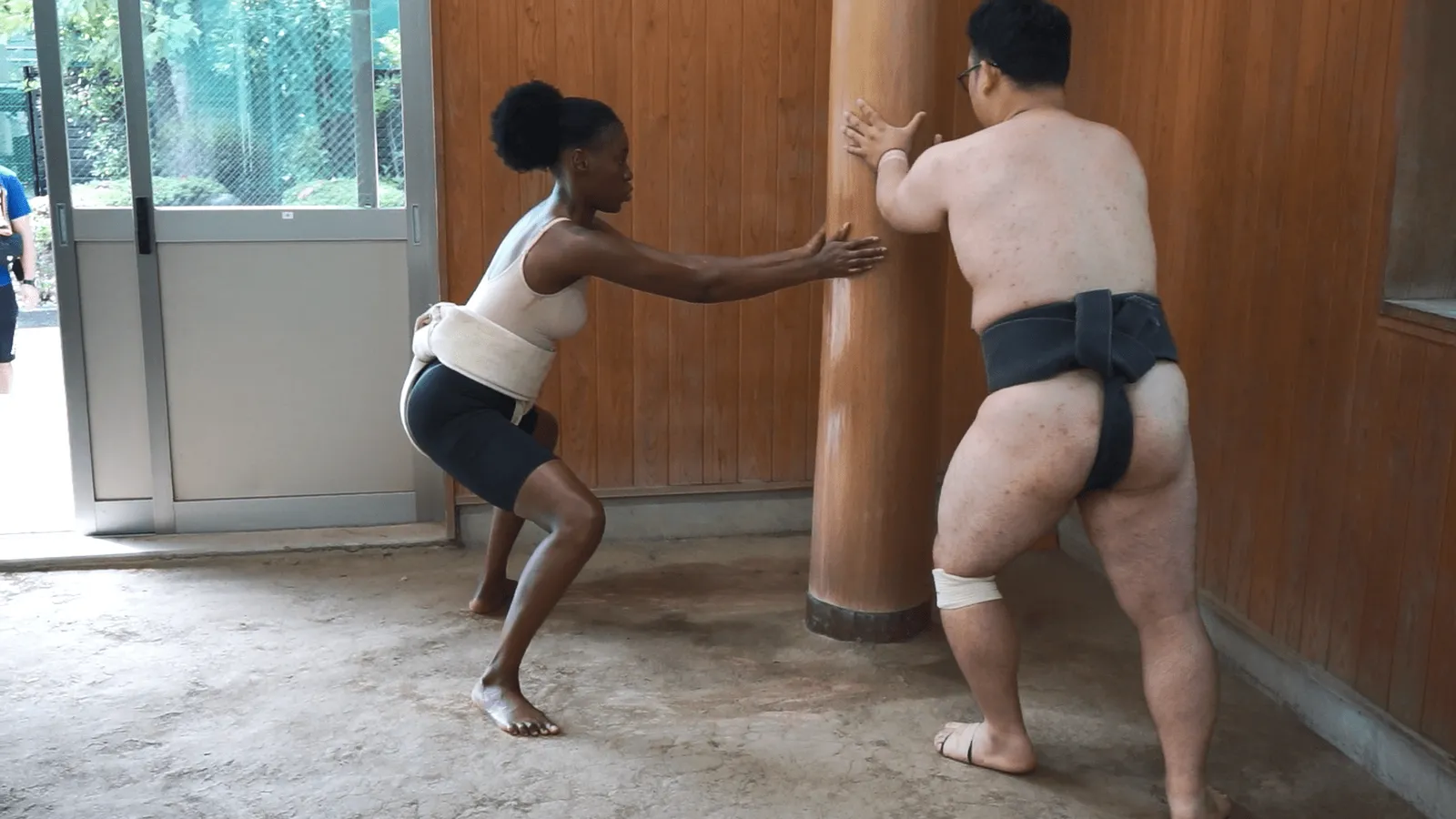 The rikishi guiding a participant through the teppo pushing motion against a post