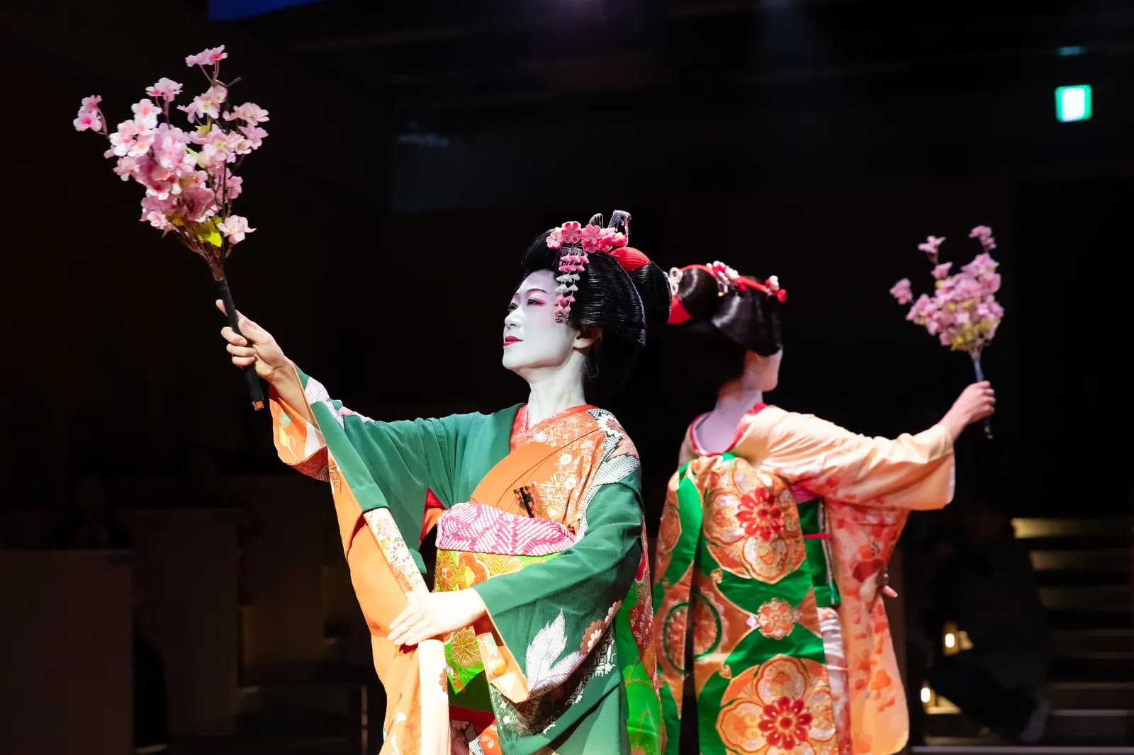 Two women in maiko-style kimono dancing while holding branches of cherry blossom