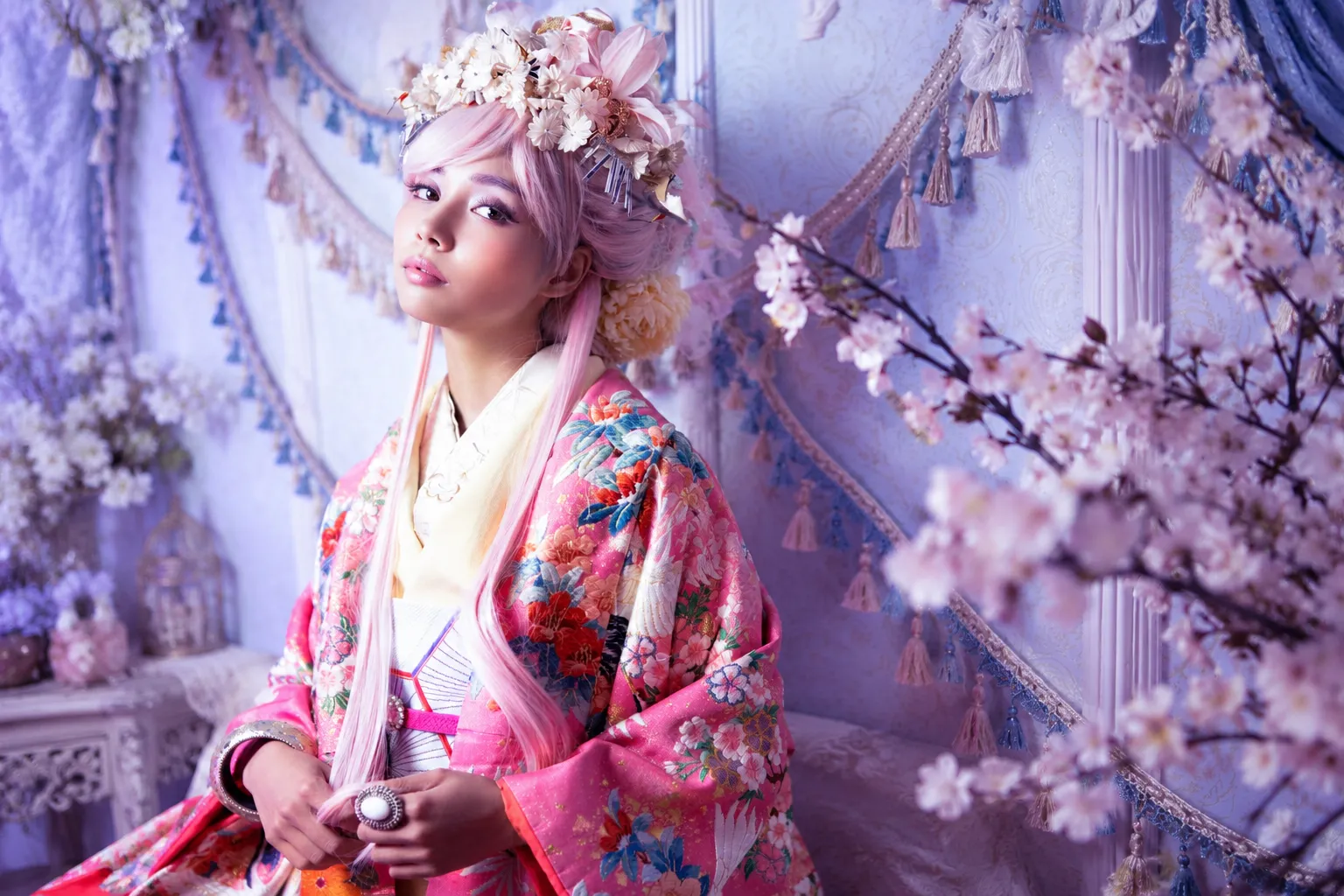 ピンクの着物と淡いピンクのウィッグ、花冠をつけ桜の前に座る女性