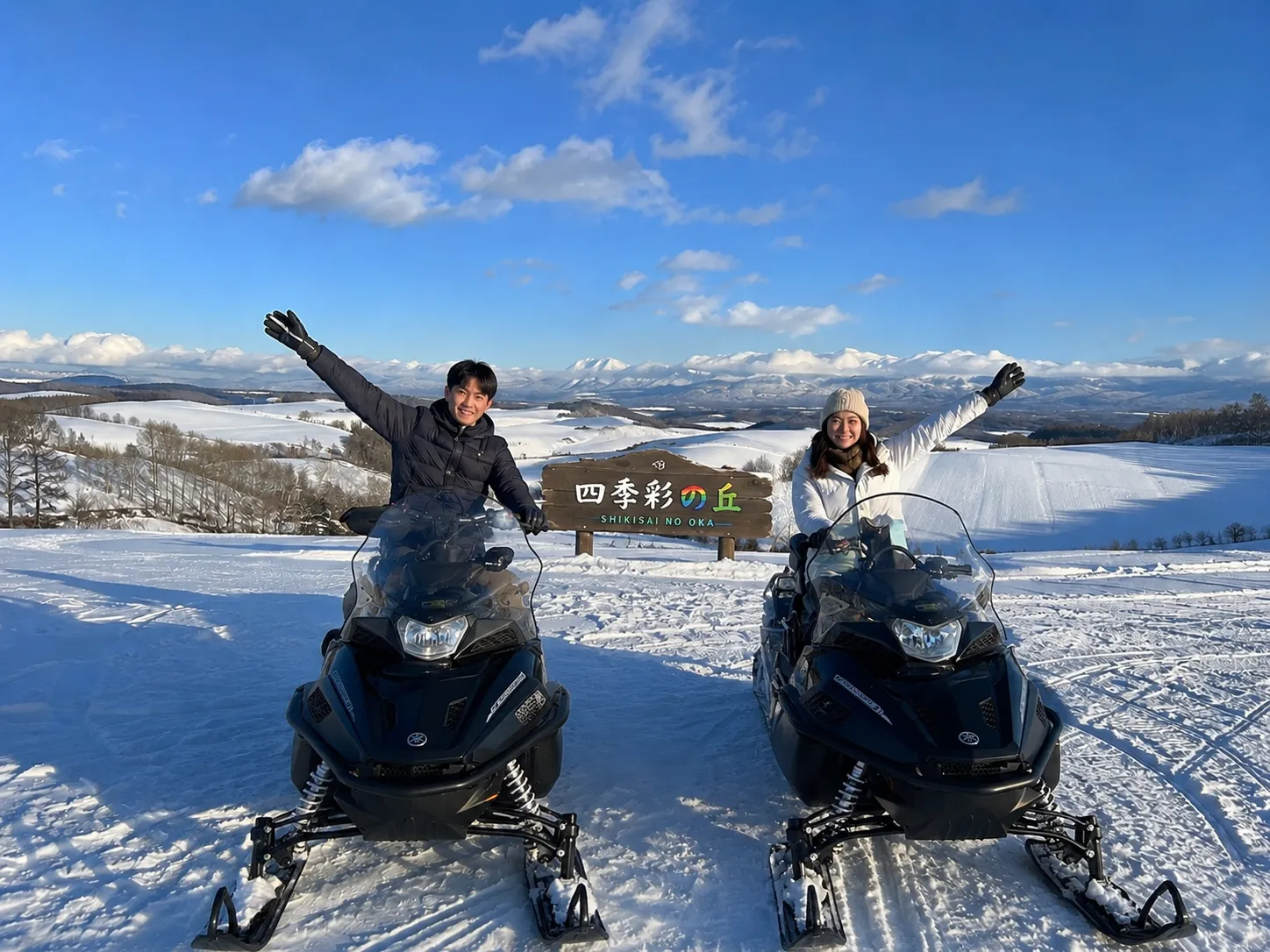 Two people on a snowmobile in front of the Shikisai no Oka sign
