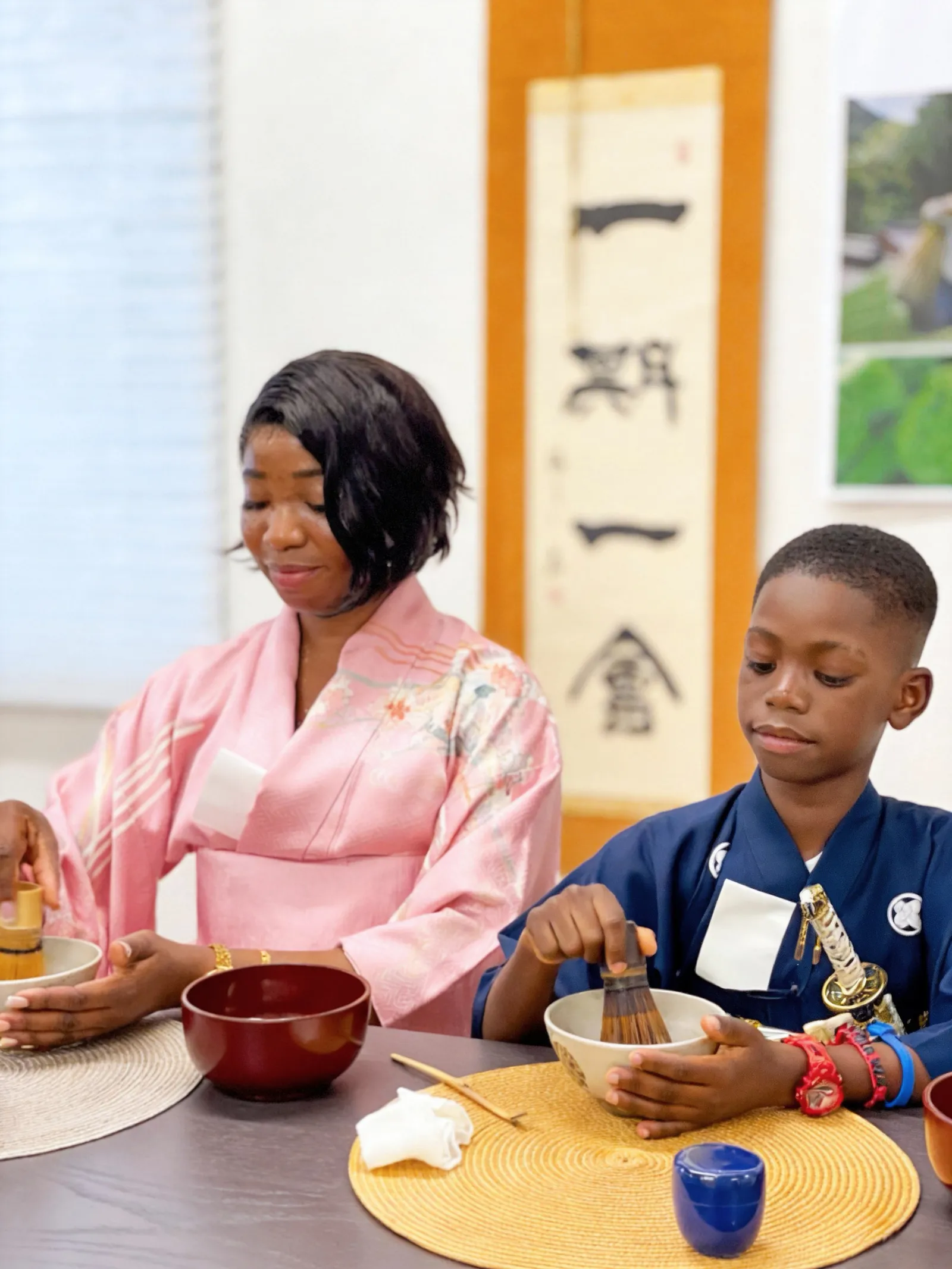 A woman in a pink kimono and a boy in a navy kimono whisking matcha with bamboo whisks