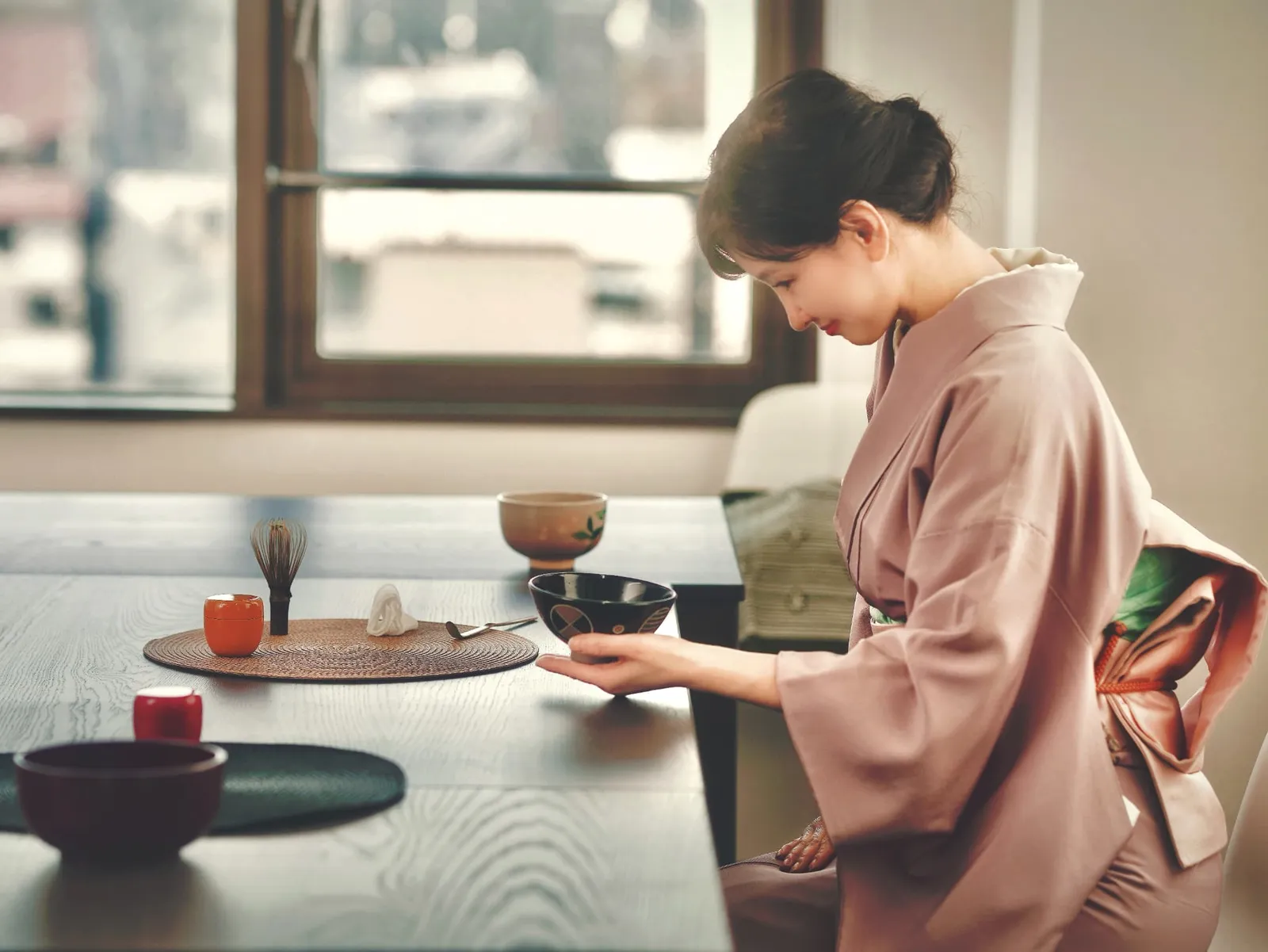 A woman in kimono holding a black tea bowl in a room with tea utensils arranged on a table