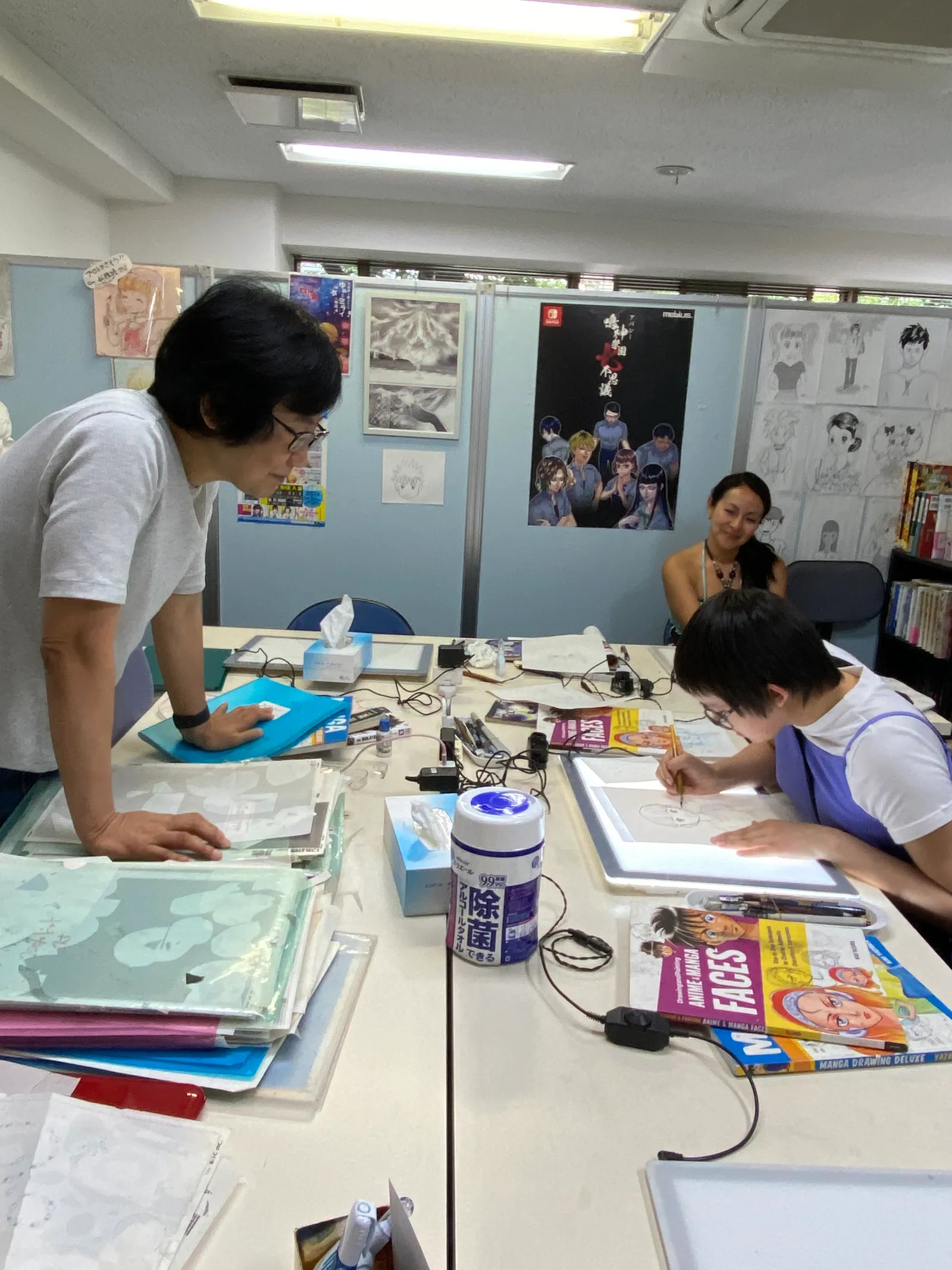 A classroom where an instructor leans on the desk and watches a participant draw on a light board
