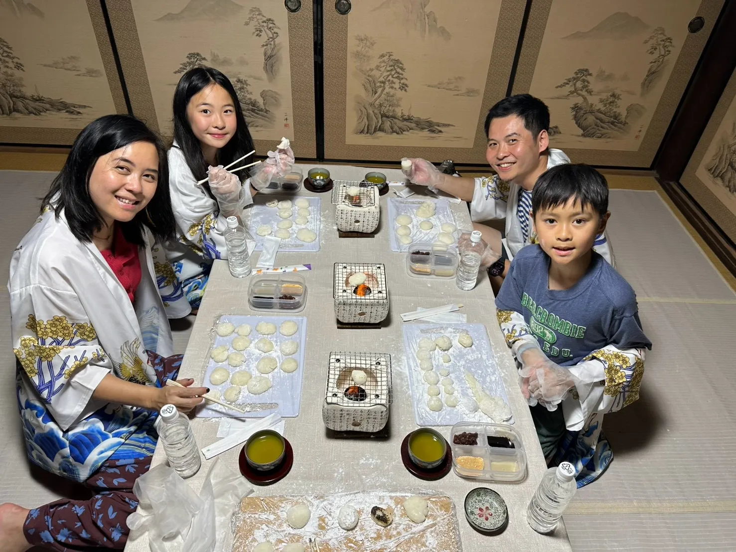 羽織を着た家族4人がまな板の上に丸めた餅を並べてテーブルを囲む様子
