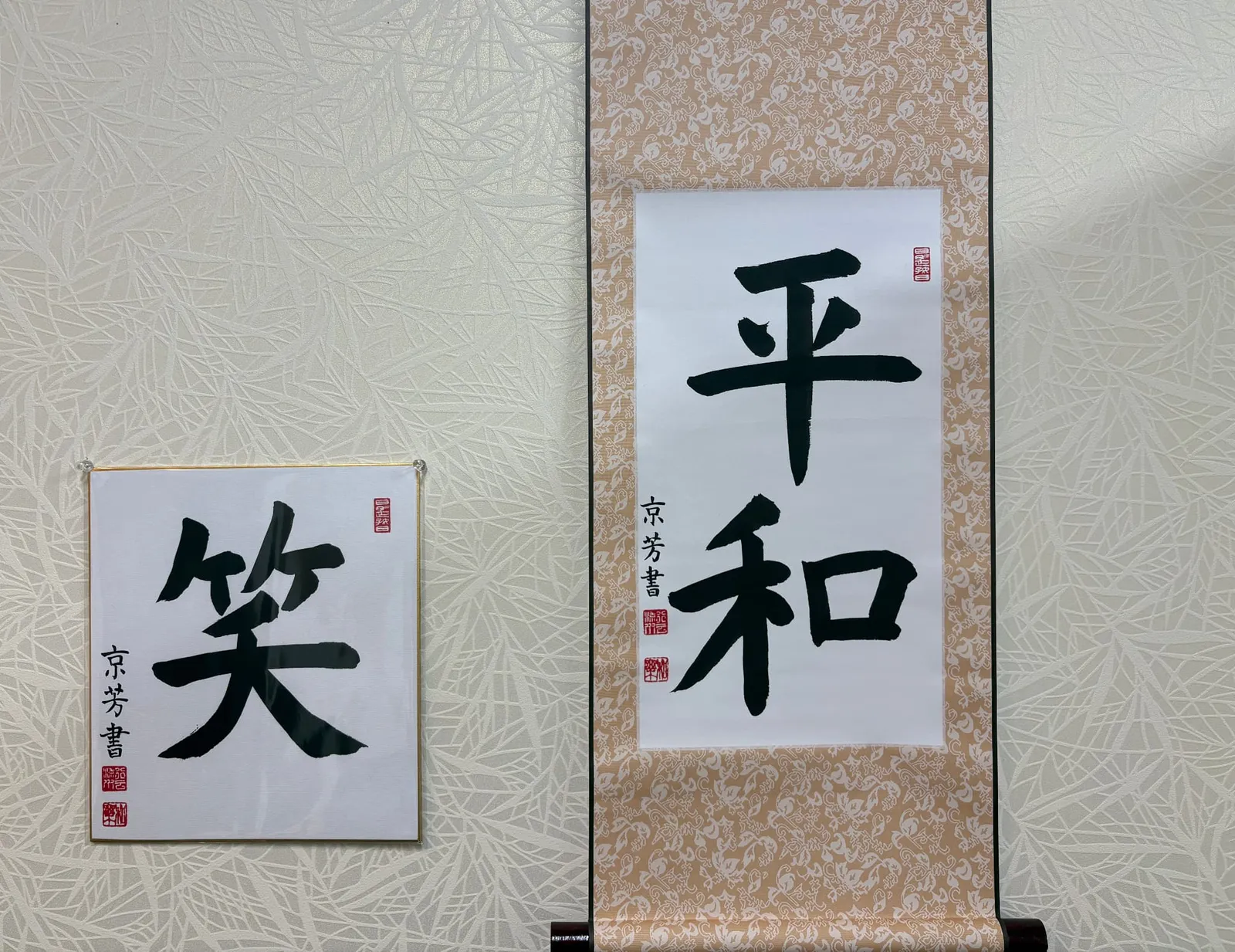 A shikishi board with the character 笑 (smile) and a hanging scroll with 平和 (peace) displayed on a wall