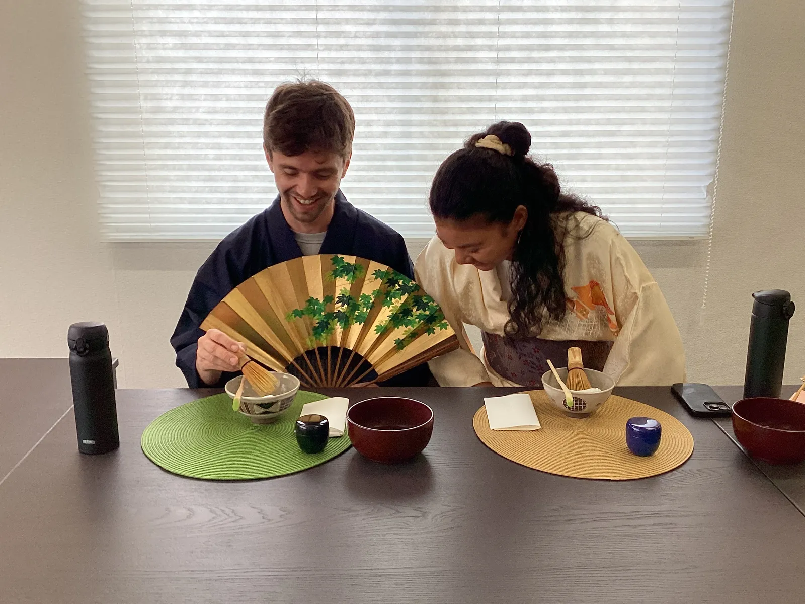 A man and a woman in kimono, each holding a folding fan and facing a tea bowl on their own mat