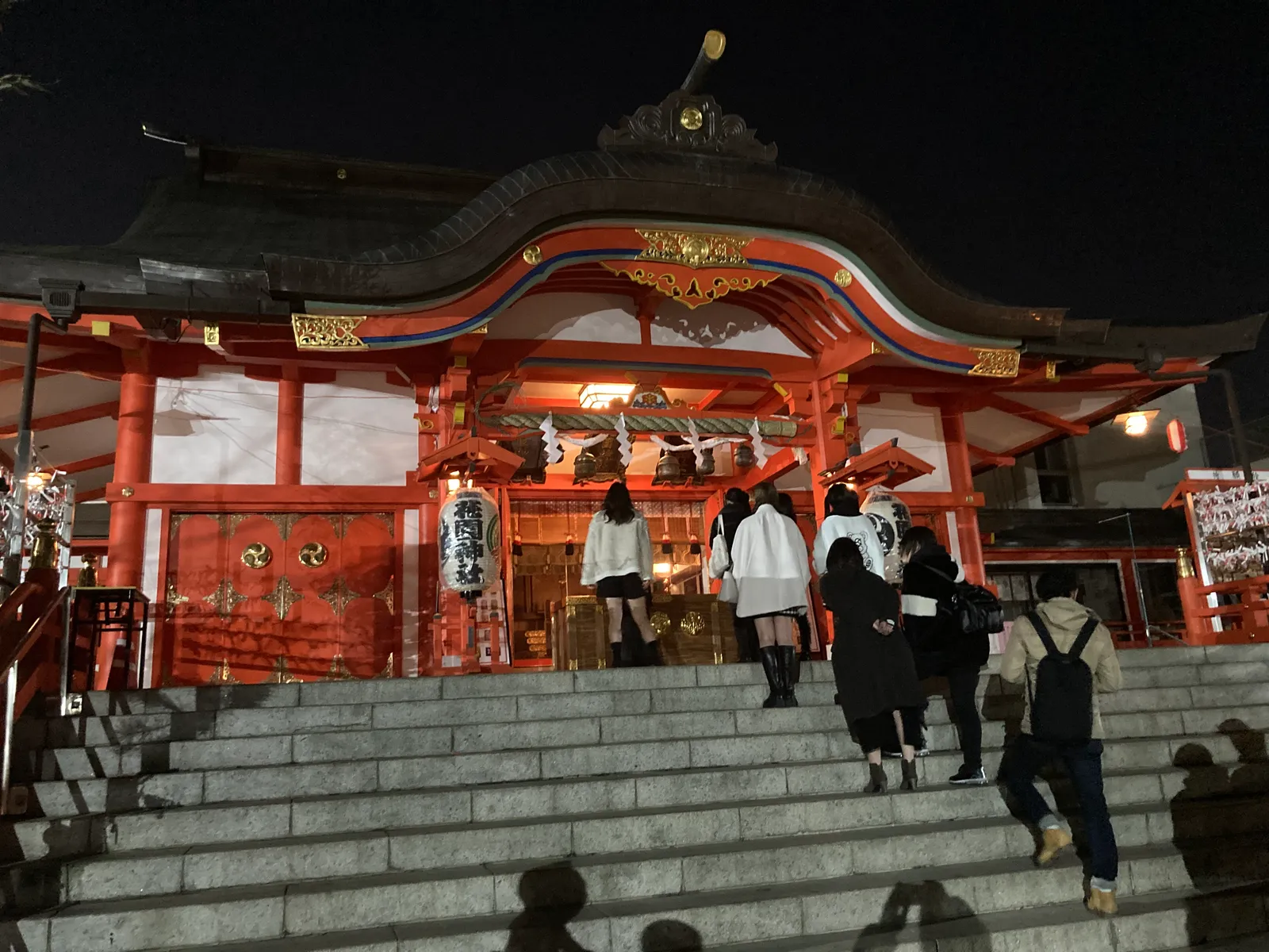 夜間にライトアップされた朱塗りの神社社殿と階段を上る参拝者