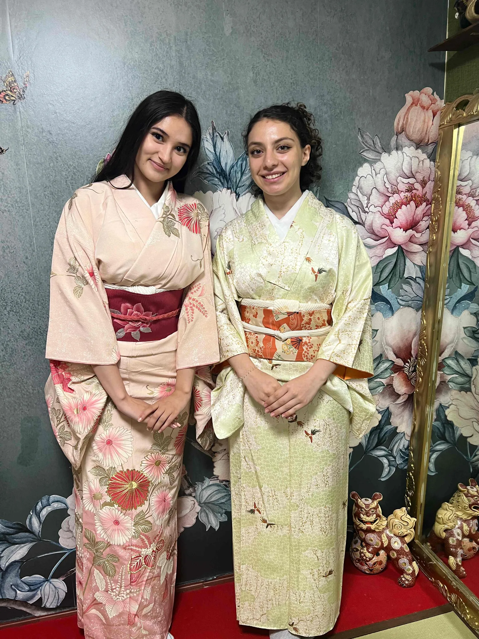 淡い色の着物を着た2人が花柄の壁の前で立つ