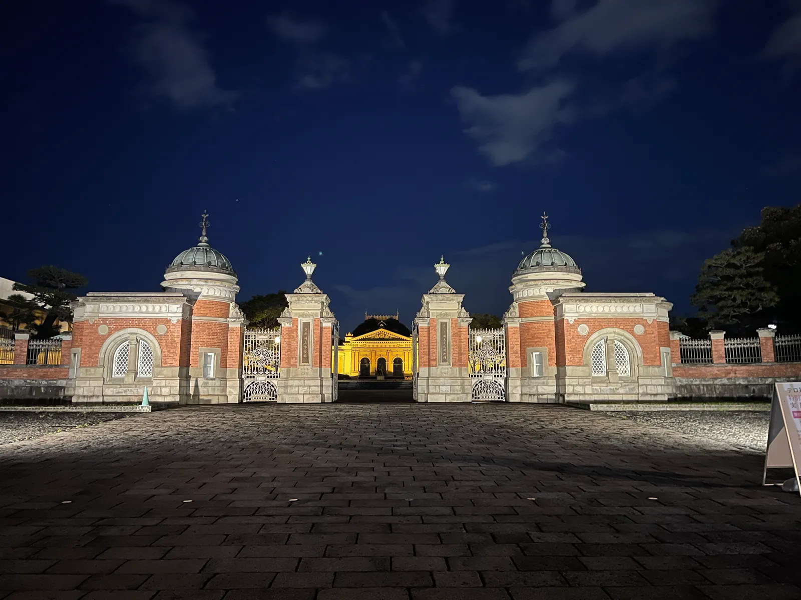 An illuminated red-brick gate at night with a Western-style building behind it
