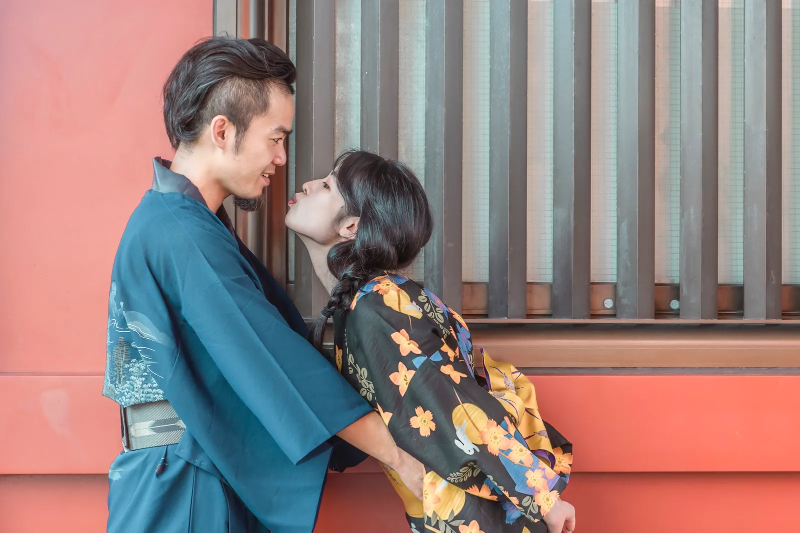 A couple in kimono gazing at each other, faces close, in front of a vermilion wall and lattice window